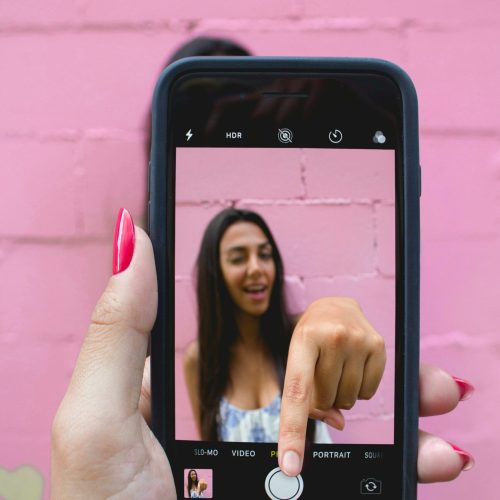 A woman captures her selfie using a smartphone against a vibrant pink wall.